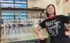 Fig. 3: Allie proudly posing in front of the stir rack that she built at FHL. Larvae had just been added to their jars for acclimation, Jan. 2023. Image credit: Fiona Curliss.