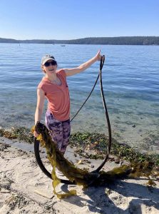 Becca Maher stands at the water's edge on a sunny day holding up a dark brown bull kelp specimen which had washed up, just for fun.